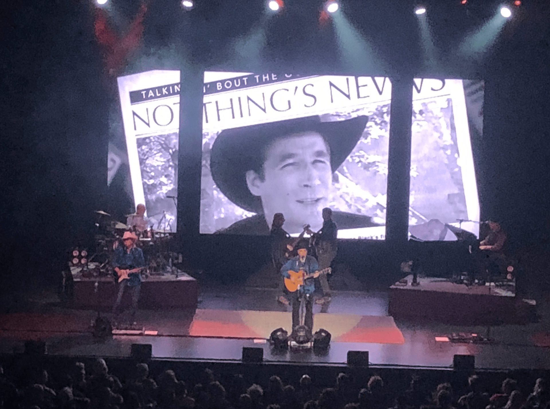 Musician performing on stage with a guitar, accompanied by band members. A large screen displays a nostalgic black-and-white photo and text.