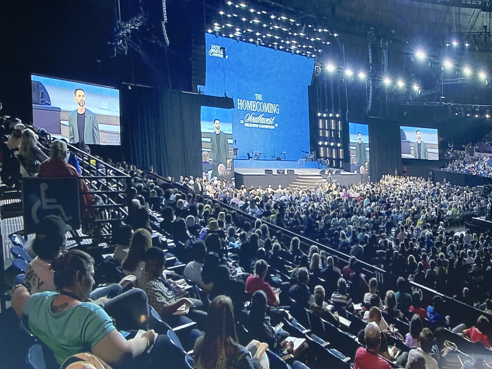 A large audience attends a speaking event titled "The Homecoming Outreach." A speaker addresses the crowd on stage, with large screens displaying visuals.