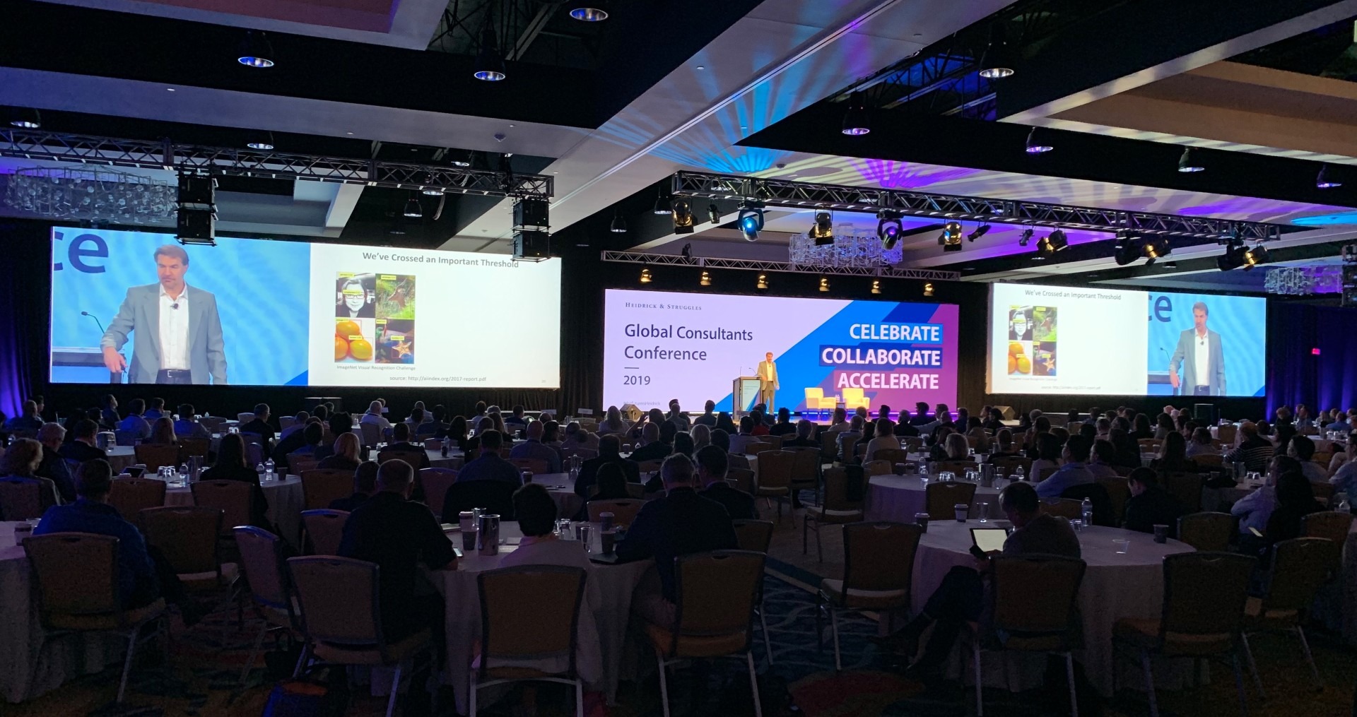 A packed global consultants conference shows a speaker on stage, colorful presentation screens, and attendees seated in a dim, energetic ballroom.